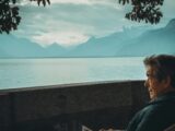 Ein Mann sitzt am Ufer eines Sees und blickt auf das Wasser und die dahinterliegenden Berge.